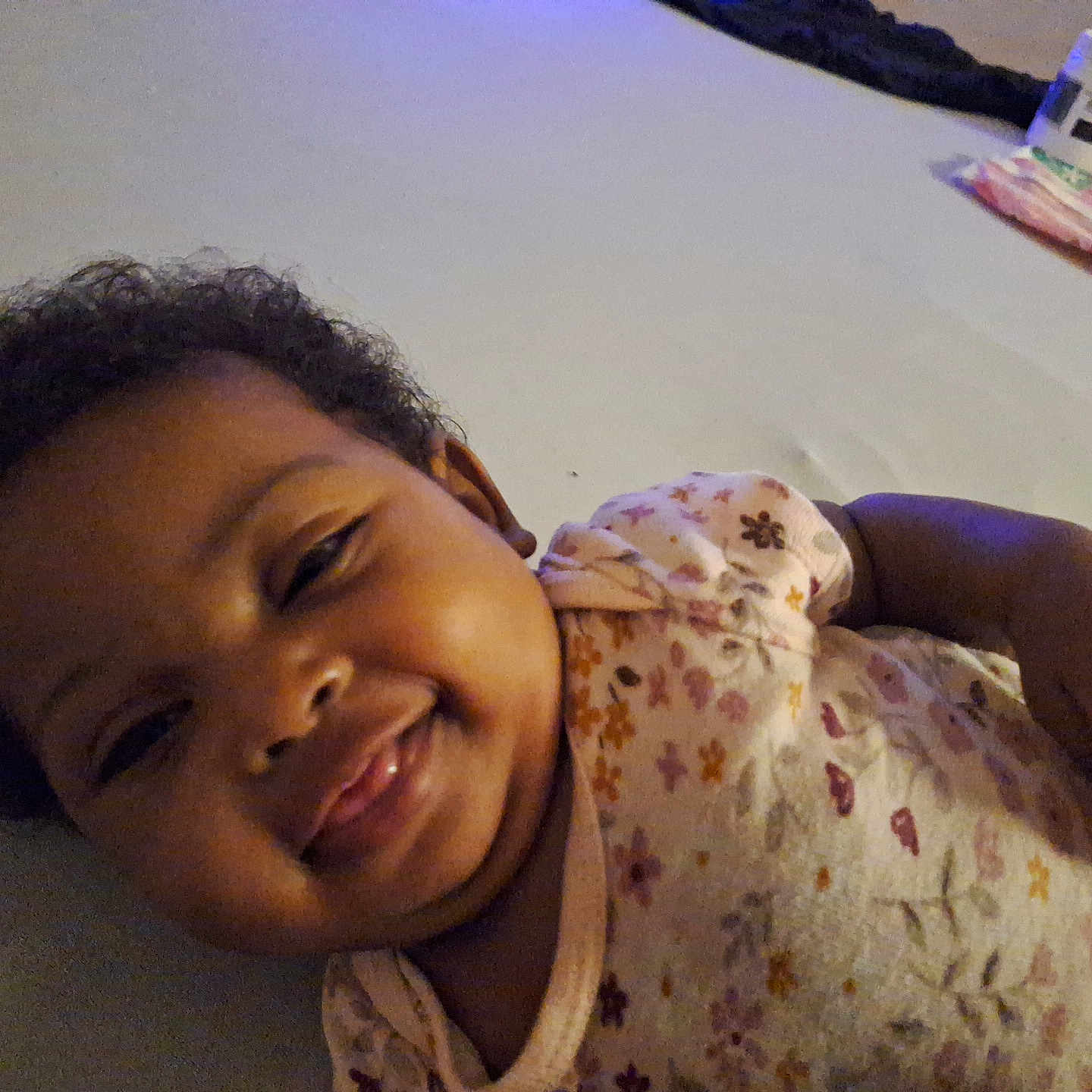 Kamari is registered to the contest to win money with this photo: baby, bed, child, comfort, cozy, curly_hair, cute, face, floral_onesie, hand, happy, indoor, infant, lying_down, person, portrait, skin, sleepwear, smiling, young_child