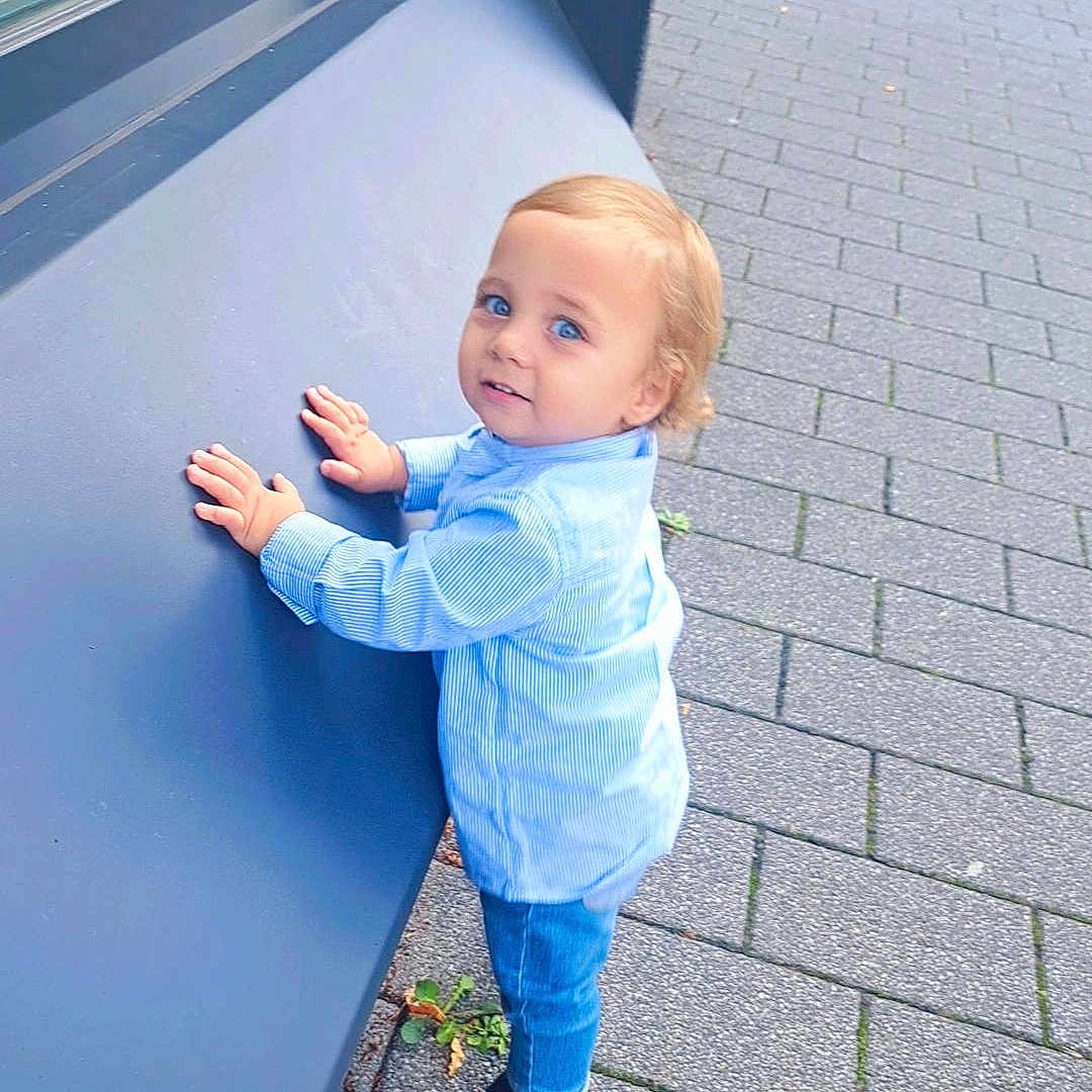 Alex participe au concours pour gagner de l'argent avec cette photo : blond_hair, blue_eyes, boy, casual_clothing, child, curious, daylight, greenery, hands_on_surface, jeans, outdoor, pavement, sandals, sidewalk, standing, striped_shirt, toddler, urban, window_reflection, young_child