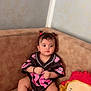baby, brown_furniture, casual_clothing, child, comfort, couch, curly_hair, cute, expression, home, indoor, infant, jersey, person, pigtails, playful, plush_doll, portrait, sitting, toy