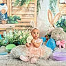 baby, child, pink_dress, bunny_ears, plush_toy, easter_eggs, flowers, cushion, carpet, indoor, decorations, celebration, spring, cute, portrait, sitting, person, holiday, festive, soft_toys