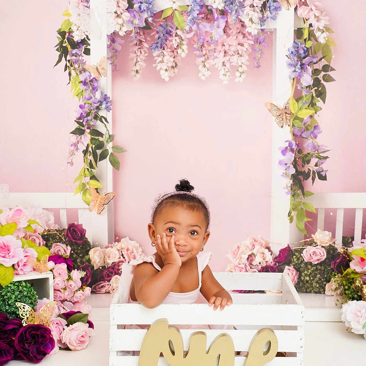 Ny'Ari is registered to the contest to win money with this photo: toddler, child, crate, flowers, floral_arch, butterflies, pink_background, portrait, cute, decor, baby, smiling, headband, white_clothing, wooden_crate, indoor, celebration, one, greenery, playful