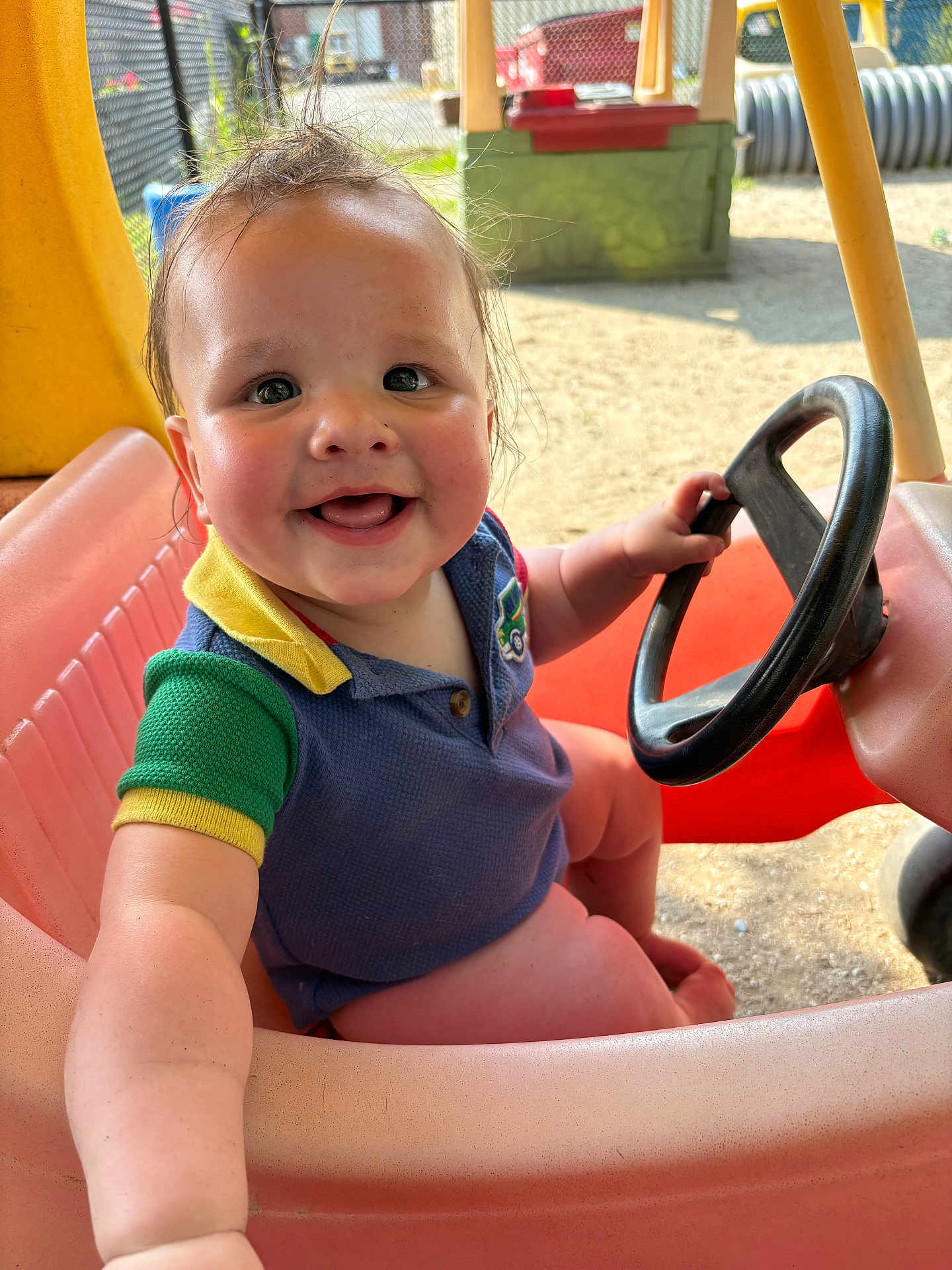 Wesley is registered to the contest to win money with this photo: baby, child, smiling, toy_car, steering_wheel, outdoor, playground, happy, cute, toddler, short_sleeve, colorful_clothing, seat, sunlight, face, hand, hair, skin, play, vehicle