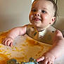 Wesley is registered to the contest to win money with this photo: baby, high_chair, bib, baby_food, messy, teether, dinosaur_toy, child, food, cute, happy, smiling, indoor, feeding, infant, chubby_cheeks, table, hand, face, portrait