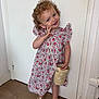 Amy participe au concours pour gagner de l'argent avec cette photo : bag, blue_eyes, child, curly_hair, cute, door, fashion, floral_dress, happy, indoors, person, playful, posing, sandals, smiling, standing, toddler, white_wall, wooden_floor, woven_bag