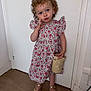 blue_eyes, child, curly_hair, cute, door, dress, fashion, floral_pattern, hand_on_face, indoors, person, playful, pose, purse, sandals, smile, standing, toddler, wooden_floor, young_child