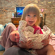 Paige is registered to the contest to win money with this photo: toddler, baby, child, playful, tongue_out, indoor, brick_wall, furniture, blanket, onesie, smiling, holding, newborn, cute, person, home, cozy, pink, shirt, portrait