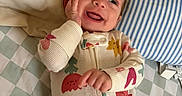 Paige is registered to the contest to win money with this photo: baby, smiling, onesie, bed, pillow, striped_pillow, checkered_sheet, infant, happy, face, hand, clothing, sleepwear, cute, lying_down, child, indoors, portrait, expression, cozy