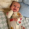 baby, smiling, onesie, bed, pillow, striped_pillow, checkered_sheet, infant, happy, face, hand, clothing, sleepwear, cute, lying_down, child, indoors, portrait, expression, cozy