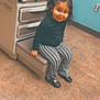 toddler, child, smile, curly_hair, black_top, houndstooth_pants, black_shoes, clinic_room, medical_equipment, floor, wall, step, sitting, cute, indoor, person, young_child, happy, healthcare, examination_table