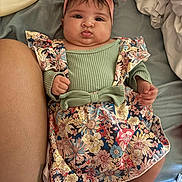 Avianna is registered to the contest to win money with this photo: baby, infant, headband, pink_bow, floral_dress, pout, lying_down, bed, blanket, lotion_bottle, skin, hand, foot, cute, child, indoors, fabric, soft, person, portrait