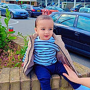 Muhammad joined the competition — help win amazing prizes! toddler, child, striped_shirt, beige_jacket, brick_wall, hand, parking_lot, cars, building, sign, sky, clouds, plant, flowers, sidewalk, person, outdoor, casual, daytime, support