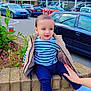 toddler, child, striped_shirt, beige_jacket, brick_wall, hand, parking_lot, cars, building, sign, sky, clouds, plant, flowers, sidewalk, person, outdoor, casual, daytime, support