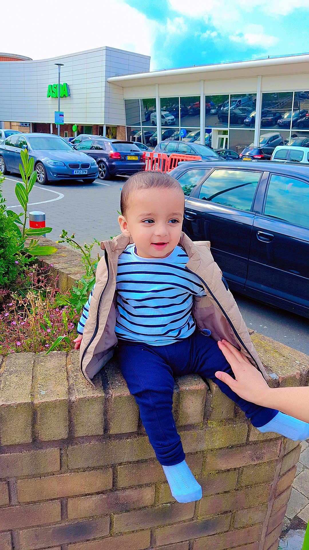 Muhammad joined the competition — help win amazing prizes! toddler, child, striped_shirt, beige_jacket, brick_wall, hand, parking_lot, cars, building, sign, sky, clouds, plant, flowers, sidewalk, person, outdoor, casual, daytime, support