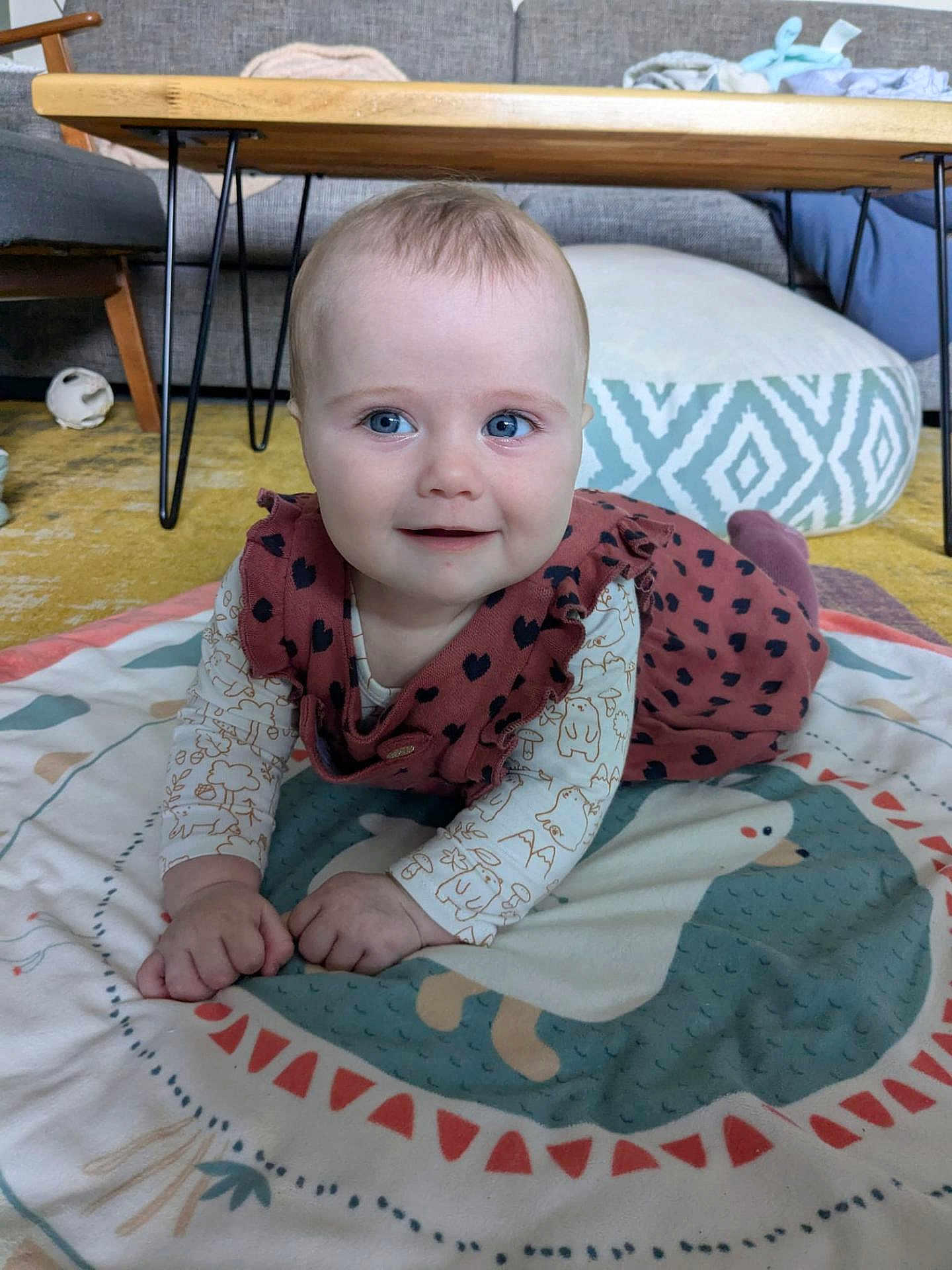 Norah participe au concours pour gagner de l'argent avec cette photo : baby, child, infant, smiling, blue_eyes, play_mat, indoor, couch, table, wooden_table, cushion, carpet, clothing, person, face, hands, cute, home, furniture, floor