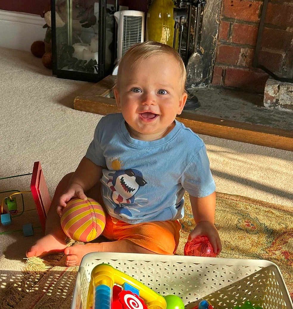 Spencer is registered to the contest to win money with this photo: toddler, child, smiling, indoor, carpet, toy, basket, colorful, fireplace, brick, shirt, orange_shorts, happy, person, foot, playtime, home, light, shadow, living_room