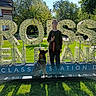 blue_sky, building, casual_clothing, daylight, decor, dog, glasses, grass, happy, leisure, nature, outdoor, park, person, shadow, signage, sitting, standing, sunlight, trees