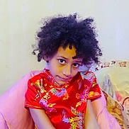 Connie is registered to the contest to win money with this photo: child, curly_hair, red_dress, floral_pattern, portrait, indoor, sitting, looking_at_camera, expressive_eyes, pink_blanket, pillow, teddy_print, couch, pose, soft_lighting, face, hands, cozy, colorful_clothing, candid