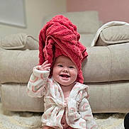 Madeline is registered to the contest to win money with this photo: baby, smiling, towel, robe, indoor, happy, child, cute, face, furniture, carpet, living_room, skin, headwear, sitting, infant, expression, clothing, comfort, playful