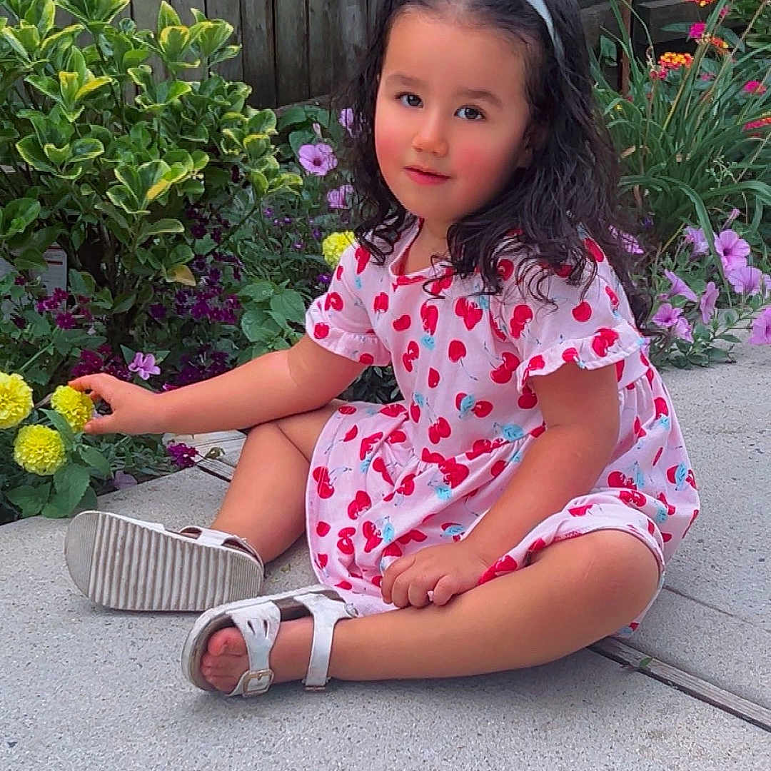 Aitana is registered to the contest to win money with this photo: cherry_pattern, child, colorful, dress, flowers, garden, girl, nature, outdoor, patio, plants, portrait, portrait_photography, sandals, sitting, smiling, toddler, white_bow, wooden_fence, yellow_flower