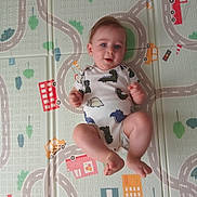 Milo joined the competition — help win amazing prizes! baby, infant, child, onesie, dinosaur_print, play_mat, colorful, roads, cars, buildings, blue_eyes, cute, lying_down, floor, hands, feet, expression, indoor, soft_lighting, playful