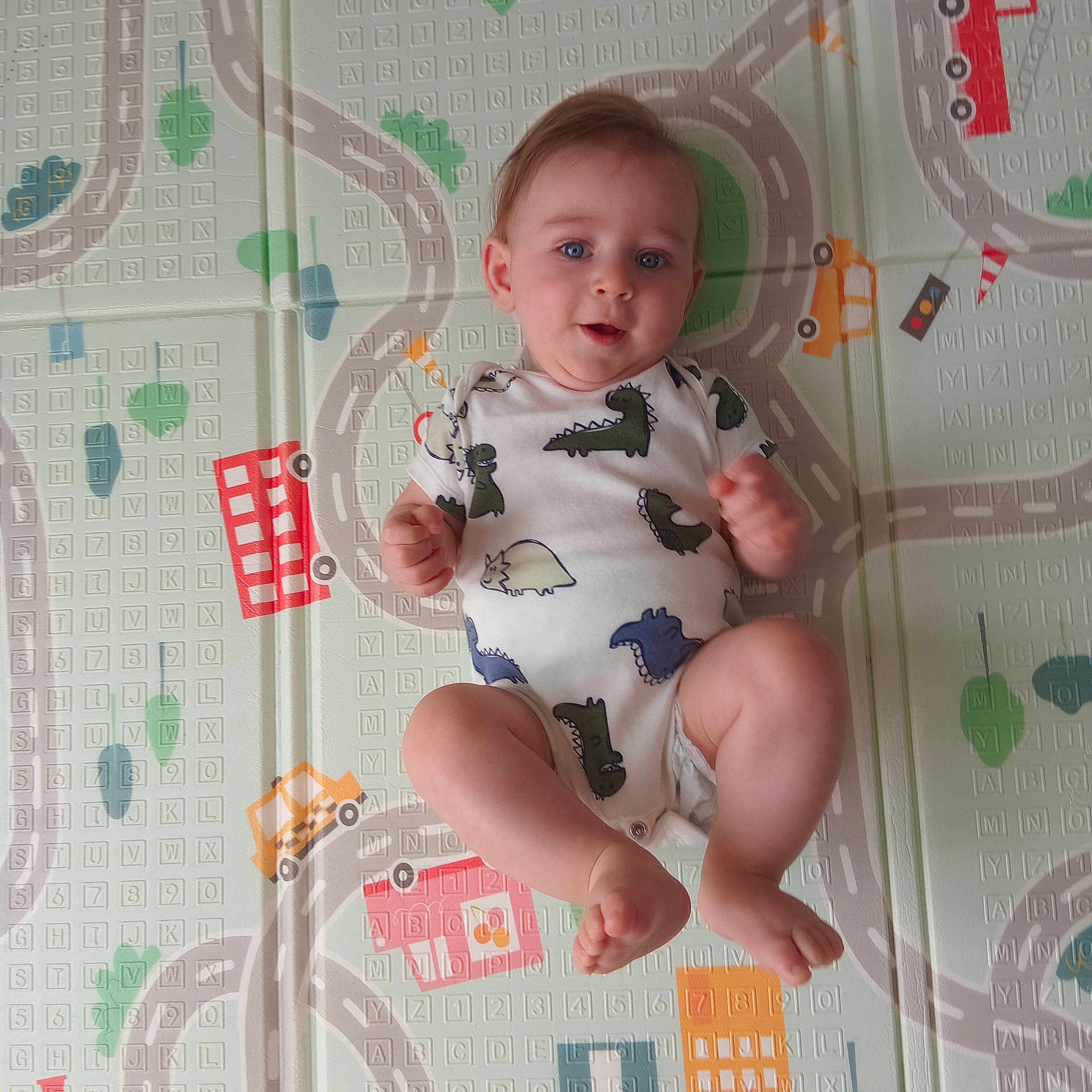 Milo joined the competition — help win amazing prizes! baby, infant, child, onesie, dinosaur_print, play_mat, colorful, roads, cars, buildings, blue_eyes, cute, lying_down, floor, hands, feet, expression, indoor, soft_lighting, playful
