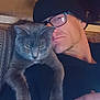 Annabelle joined the competition — help win amazing prizes! man, cat, glasses, beanie, indoor, cuddle, pet, gray_cat, closeup, portrait, face, hand, furniture, warm_lighting, cozy, relaxed, animal, person, resting, casual
