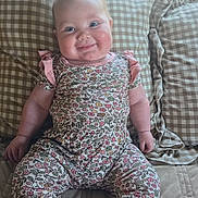 Dottie is registered to the contest to win money with this photo: baby, child, smiling, floral_outfit, pink_cuffs, bed, checkered_pillows, couch, indoors, happy, person, infant, cute, chubby_cheeks, seated, portrait, soft_lighting, cozy, home, relaxed