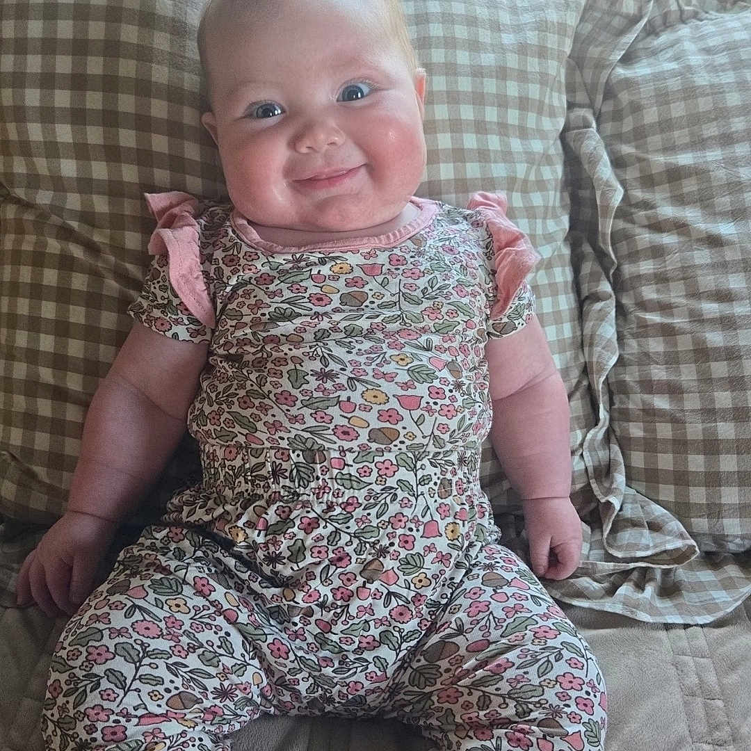 Dottie is registered to the contest to win money with this photo: baby, child, smiling, floral_outfit, pink_cuffs, bed, checkered_pillows, couch, indoors, happy, person, infant, cute, chubby_cheeks, seated, portrait, soft_lighting, cozy, home, relaxed
