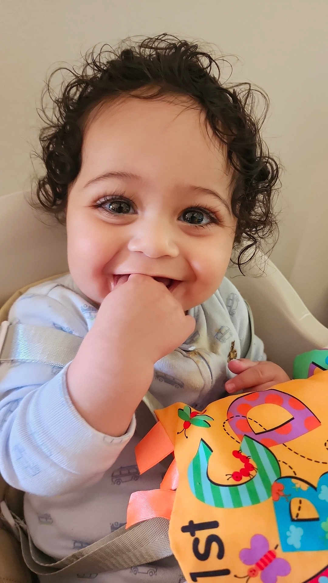 Kaveh joined the competition — help win amazing prizes! baby, infant, child, curly_hair, smiling, teeth, hand_in_mouth, big_eyes, long_eyelashes, high_chair, seat_strap, colorful_toy, fabric_book, pajamas, indoor, portrait, cute, playful, close_up, cheeky_grin