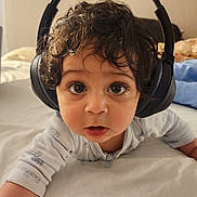 Kaveh joined the competition — help win amazing prizes! child, baby, infant, headphones, curly_hair, big_eyes, lying_down, bed, pajamas, surprised_expression, close_up, portrait, indoor, cute, audio, technology, blanket, pillow, skin, face