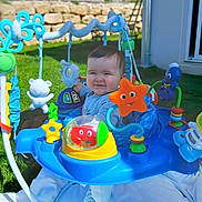Sergio a rejoint le concours — aidez-le/la à gagner de superbes lots ! baby, child, smiling, outdoor, grass, toy, activity_jumper, play, colorful, blue, orange, green, plastic, happy, infant, sunlight, face, clothing, person, garden