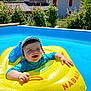 baby, child, inflatable_ring, pool, water, sun_hat, smiling, outdoor, summer, blue, yellow, house, greenery, sunny, happy, floating, swimming, vacation, playful, daytime
