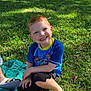 child, boy, smiling, grass, park, pizza, food, blue_shirt, red_hair, green_shoes, sunlight, outdoor, picnic, tree, leafy, casual_clothing, happy, sitting, daytime, nature