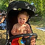 toddler, child, stroller, outdoor, sunlight, toy, pop_it, shirtless, grass, tree, summer, play, baby, nature, park, people, shade, feet, expression, casual