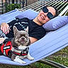 person, dog, hammock, outdoor, grass, sunglasses, smiling, leash, harness, relaxing, casual_clothing, purple, sunny, pet, happy, resting, backyard, fence, nature, daytime