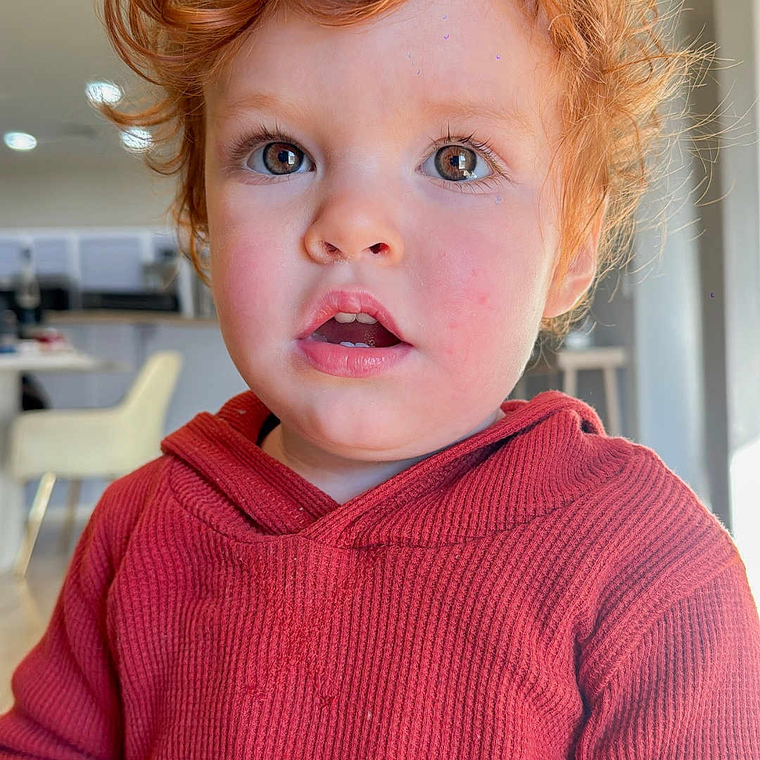 Adriel is registered to the contest to win money with this photo: chair, child, curiosity, curly_hair, expression, eyes, face, furniture, hand, hoodie, indoors, lighting, person, portrait, red_clothing, red_hair, skin, table, toddler, young_child