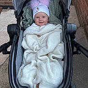 Sienna is registered to the contest to win money with this photo: baby, stroller, blanket, hat, pom_poms, smile, outdoor, sidewalk, brick_wall, clothing, child, cold_weather, happy, person, cozy, wrapped, infant, cute, transport, cute_accessory