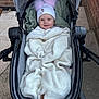 baby, stroller, blanket, hat, pom_poms, smile, outdoor, sidewalk, brick_wall, clothing, child, cold_weather, happy, person, cozy, wrapped, infant, cute, transport, cute_accessory