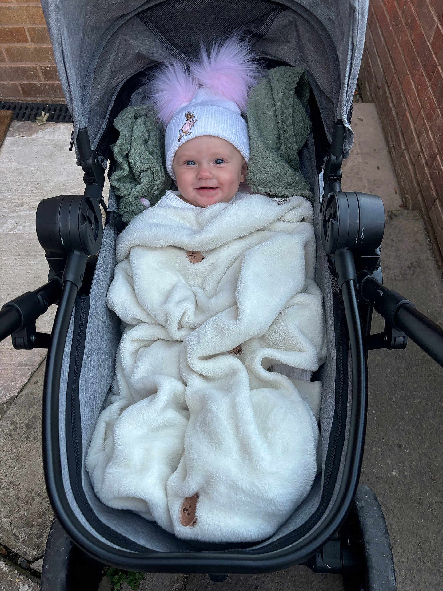 Sienna is registered to the contest to win money with this photo: baby, stroller, blanket, hat, pom_poms, smile, outdoor, sidewalk, brick_wall, clothing, child, cold_weather, happy, person, cozy, wrapped, infant, cute, transport, cute_accessory
