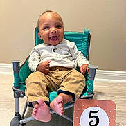 Maliki joined the competition — help win amazing prizes! baby, infant, smiling, chair, teal_chair, milestone_card, five_months, feet, toes, onesie, pants, happy, face, eyes, tongue, indoor, wooden_floor, wall, portrait, sitting