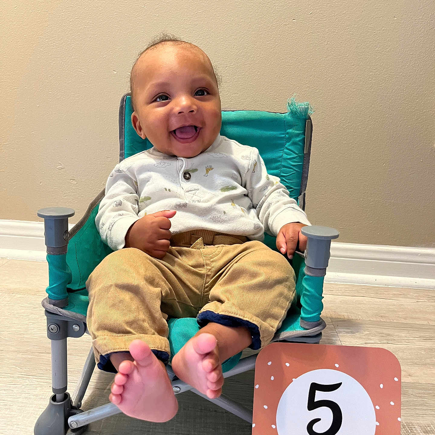 Maliki joined the competition — help win amazing prizes! baby, chair, eyes, face, feet, five_months, happy, indoor, infant, milestone_card, onesie, pants, portrait, sitting, smiling, teal_chair, toes, tongue, wall, wooden_floor