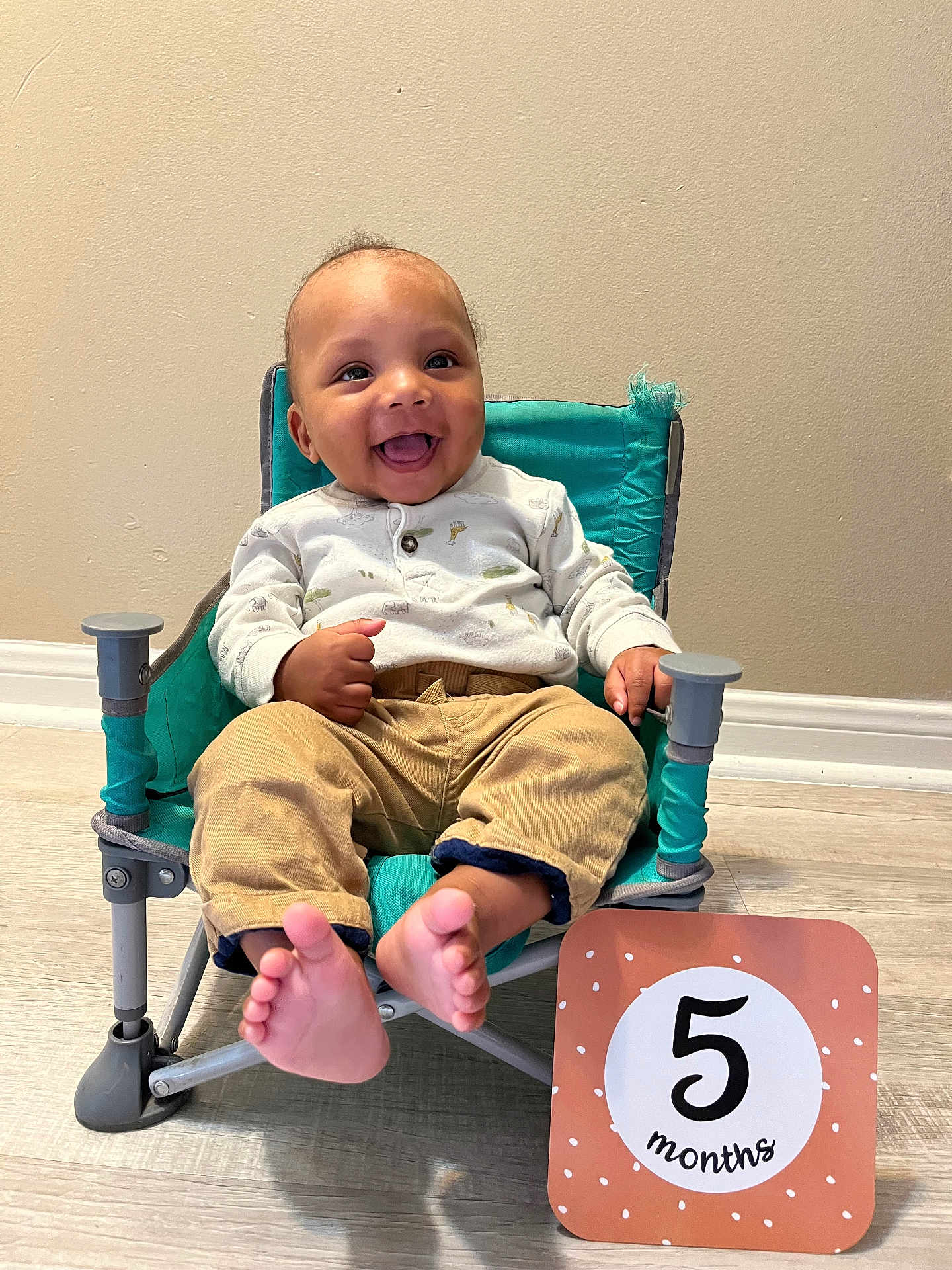 Maliki joined the competition — help win amazing prizes! baby, infant, smiling, chair, teal_chair, milestone_card, five_months, feet, toes, onesie, pants, happy, face, eyes, tongue, indoor, wooden_floor, wall, portrait, sitting