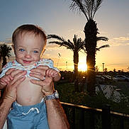 Theodore joined the competition — help win amazing prizes! baby, child, smile, hands, sunset, palm_trees, blue_sky, outdoor, person, cute, happy, portrait, shorts, shirt, nature, sunlight, parking_lot, fence, adult, holding