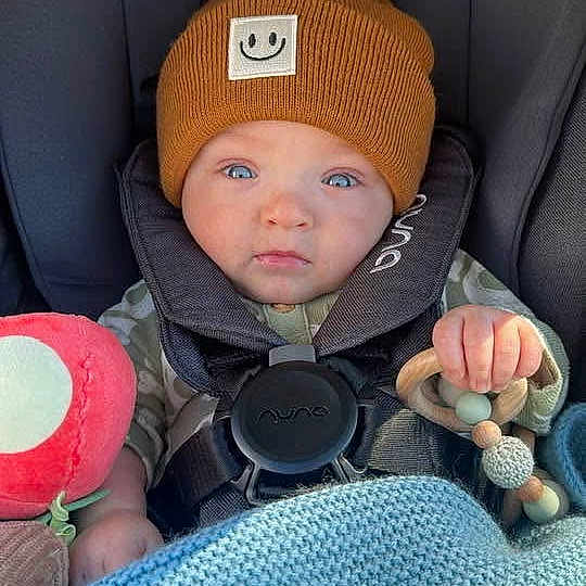 Theodore is registered to the contest to win money with this photo: baby, car_seat, beanie, blue_blanket, wooden_toy, face, child, infant, clothing, seatbelt, cute, indoors, portrait, expression, hand, knit_blanket, smile_patch, looking, sitting, warm