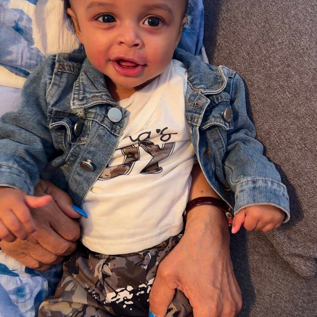 Ashad is registered to the contest to win money with this photo: adult_hand, blue_nail_polish, casual_clothing, child, close_up, couch, curly_hair, cute, denim_jacket, family, happy, holding, indoor, patterned_pants, person, portrait, seated, smiling, toddler, young_child