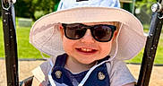 Cameron is registered to the contest to win money with this photo: baby, child, clothing, daylight, face, fun, happy, hat, outdoor, park, person, playground, seat, smile, summer, sunglasses, sunhat, sunny, swing, toddler