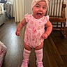 baby, child, girl, pink_clothing, headband, bow, smiling, happy, standing, shoes, socks, wooden_floor, rocking_chair, indoor, room, furniture, cute, expression, person, portrait
