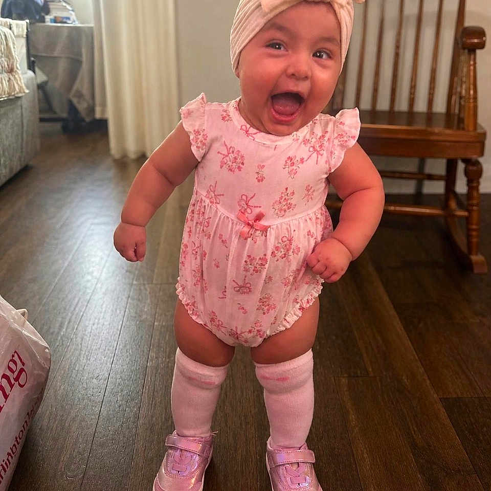 Galilea Ocon joined the competition — help win amazing prizes! baby, child, girl, pink_clothing, headband, bow, smiling, happy, standing, shoes, socks, wooden_floor, rocking_chair, indoor, room, furniture, cute, expression, person, portrait