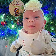 Harper is registered to the contest to win money with this photo: baby, child, headband, mittens, christmas_tree, ornament, holiday, festive, lights, face, person, winter_clothing, indoors, cute, celebration, holiday_decor, smiling, young_child, christmas, seasonal