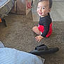 toddler, child, smiling, carpet, slipper, pillow, paper_bag, door, floor, indoor, clothing, person, home, happy, sitting, black_shirt, red_shorts, head, face, light